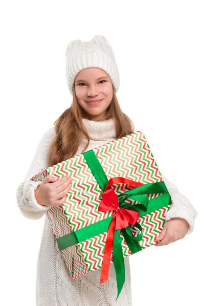 Retrato do comprimento total da menina criança bonitinha no elegante suéter de malha com presente, olhando para a câmera e sorrindo, em pé contra a parede branca do estúdio. Conceito de moda infantil