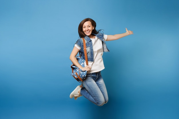 Retrato do comprimento total do estudante jovem feliz alegre em roupas jeans com mochila pulando e espalhando as mãos isoladas sobre fundo azul. Educação na faculdade. Copie o espaço para anúncio.