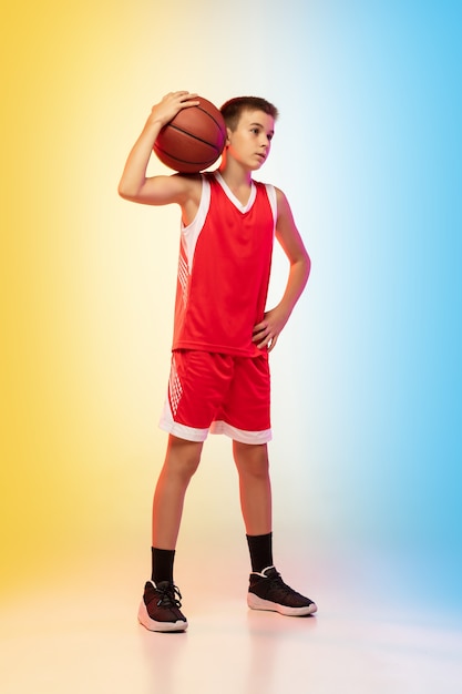 Retrato do comprimento total do jovem jogador de basquete de uniforme em fundo gradiente de estúdio. adolescente confiante posando com bola. conceito de esporte, movimento, estilo de vida saudável, anúncio, ação, movimento.