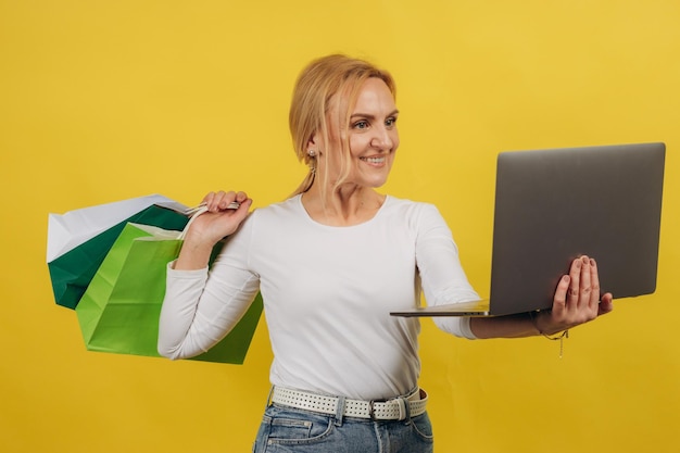 Retrato do conceito de compras on-line de uma mulher madura alegre feliz com laptop segurando sacolas de compras isoladas no fundo amarelo do estúdio Mulher excitada desfrutando de desconto