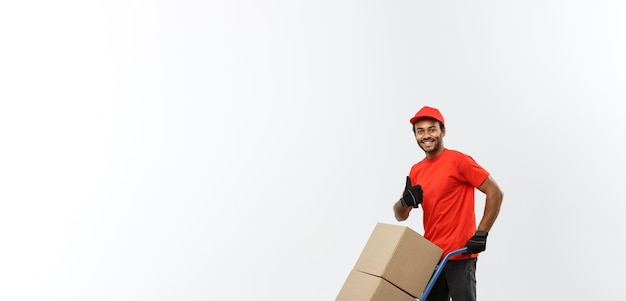 Retrato do conceito de entrega de um entregador afro-americano bonito ou mensageiro empurrando o caminhão de mão com pilha de caixas isoladas no espaço de cópia de fundo do estúdio cinza