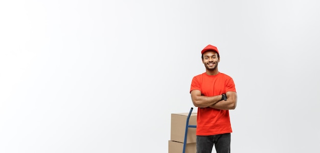 Retrato do conceito de entrega de um entregador afro-americano bonito ou mensageiro empurrando o caminhão de mão com pilha de caixas isoladas no espaço de cópia de fundo do estúdio cinza