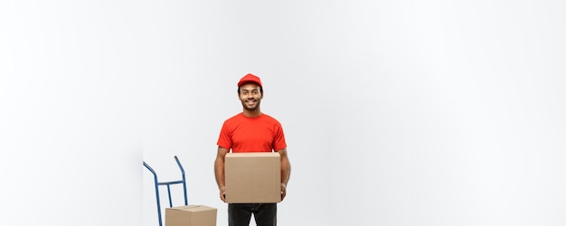 Retrato do conceito de entrega de um entregador ou mensageiro afro-americano bonito com carrinho de mão e caixa de retenção isolada no espaço de cópia de fundo do estúdio cinza