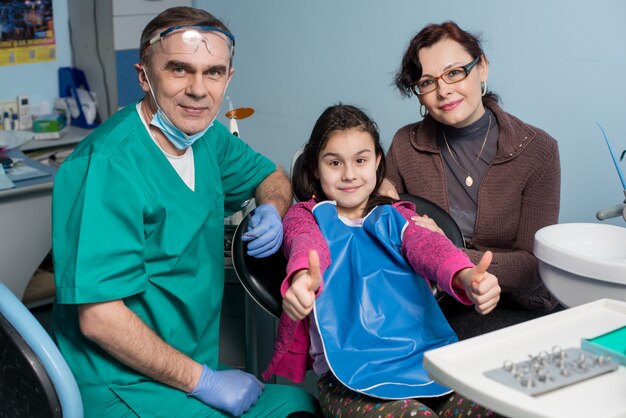 Retrato do dentista sênior e jovem garota com sua mãe na primeira visita odontológica no consultório odontológico.