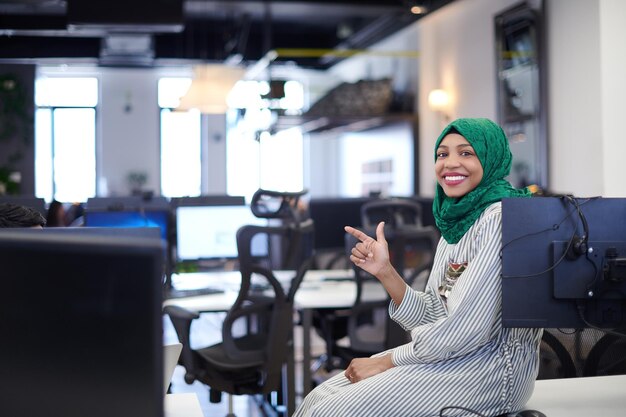 Retrato do desenvolvedor de software feminino africano muçulmano com hijab verde sentado na mesa no escritório de inicialização de plano aberto moderno