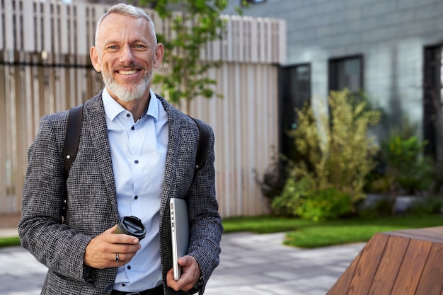 Foto retrato do elegante empresário de meia-idade, sorrindo para a câmera enquanto caminhava com seu laptop e um