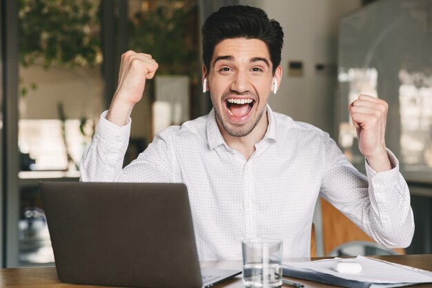 Retrato do empresário alegre dos anos 30, vestindo camisa branca e fones de ouvido sem fio, gritando e cerrando os punhos como vencedor, enquanto está sentado no escritório