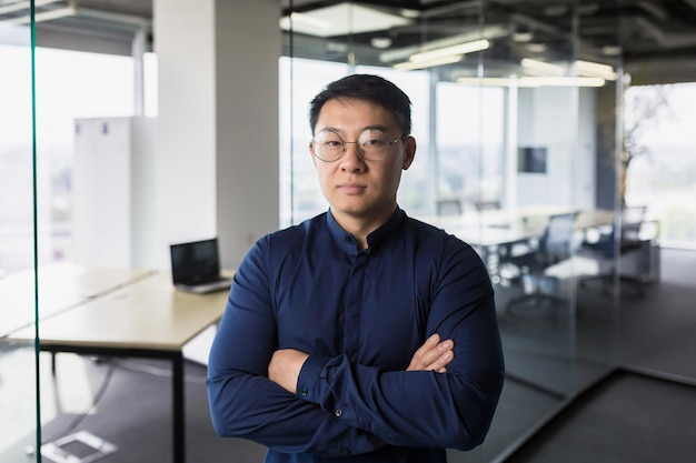 Retrato do empresário asiático dentro do chefe do escritório moderno em óculos olhando sério para o homem da câmera