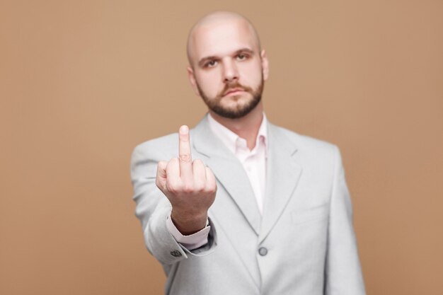 Retrato do empresário barbudo careca de meia idade bonito sério no clássico terno cinza claro em pé olhando para a câmera e mostrando o dedo médio com cara de pôquer. foto do estúdio em fundo marrom claro