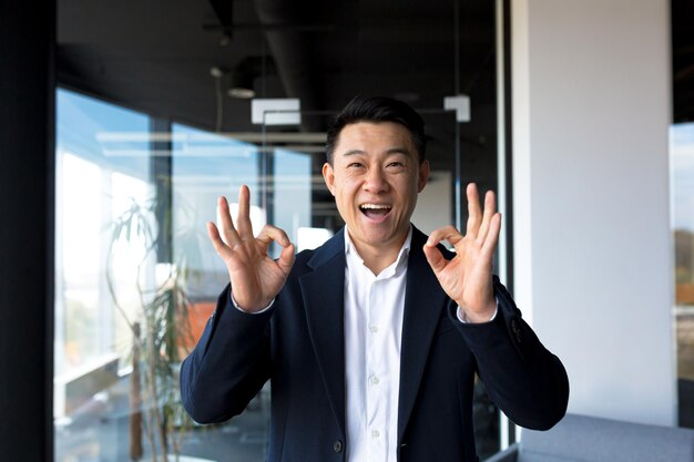 Retrato do empresário bem sucedido chefe masculino asiático alegre feliz sorrindo e regozijando-se no escritório moderno pela janela grande