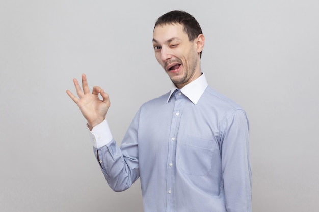 Retrato do empresário de cerdas bonito louco engraçado na camisa azul clara clássica em pé com sinal de ok e olhando para a câmera, piscando. tiro de estúdio interior, isolado em copyspace de fundo cinza.