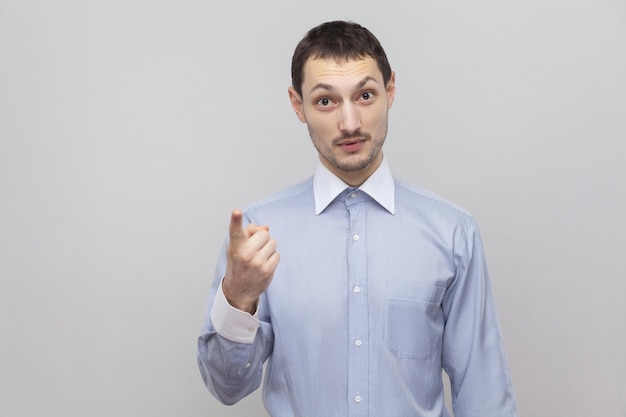 Retrato do empresário de cerdas bonito sério na camisa azul clara clássica em pé e olhando para a câmera com sinal de aviso. tiro de estúdio interior, isolado em copyspace de fundo cinza.