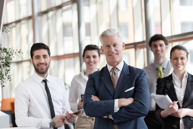 retrato do empresário sênior como líder no interior do escritório moderno e brilhante, grupo de jovens em segundo plano como equipe