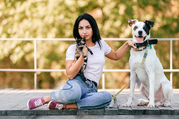 Retrato do estilo de vida da mulher morena bonita com gatinho e cachorro grande sentado ao ar livre no parque.