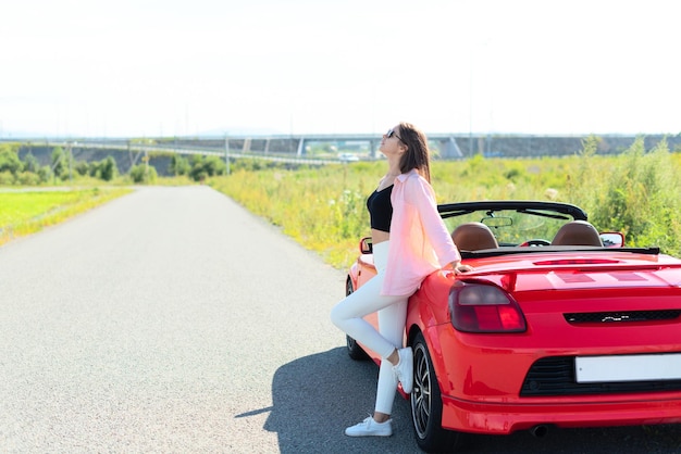 Retrato do estilo de vida de uma linda mulher despreocupada vestida casualmente em pé perto do carro cabriolet vermelho e sorrindo viagem desfrutando do conceito de liberdade