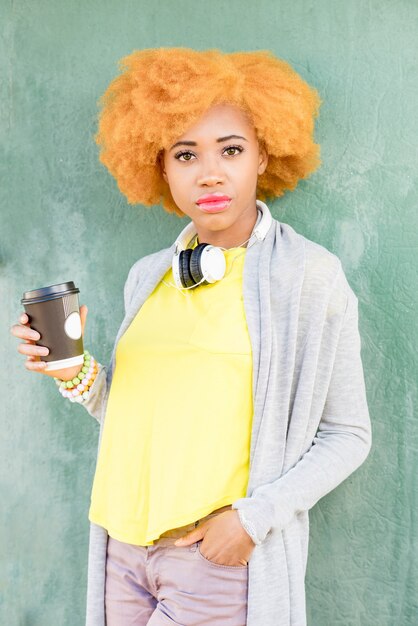 Retrato do estilo de vida de uma mulher africana em roupas casuais, em pé com café no fundo da parede verde.