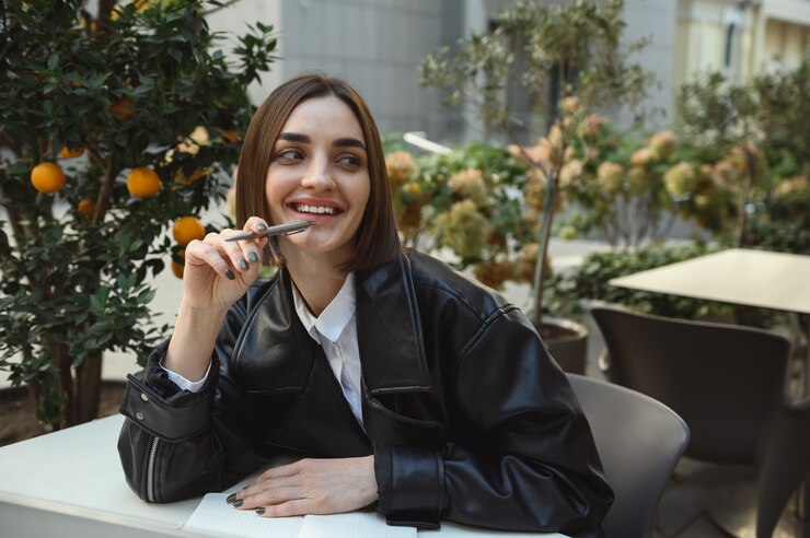 Foto retrato do estilo de vida de uma mulher bonita morena atraente elegante em uma jaqueta de couro ecológica, segurando uma caneta perto dos lábios, sorrindo, olhando de lado, sentado no terraço do café, no contexto de árvores de tangerina
