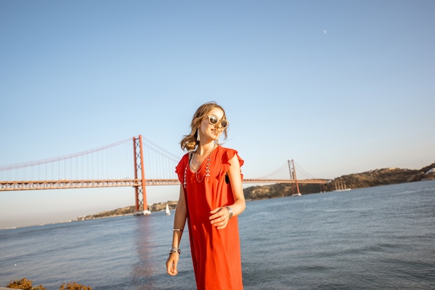 Retrato do estilo de vida de uma mulher vestida de vermelho caminhando à beira do rio com uma bela ponte de ferro ao fundo na cidade de Lisboa, Porugal