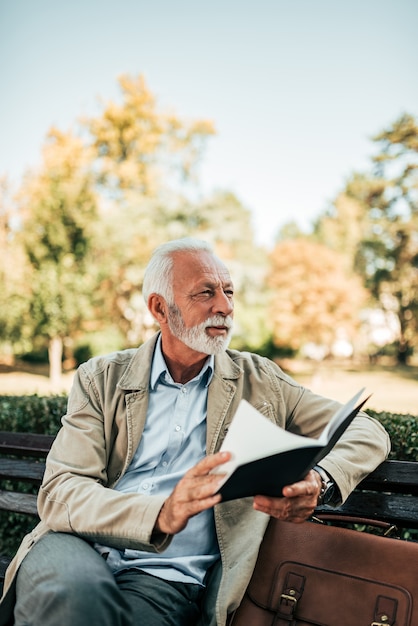 Retrato do homem aposentado que lê um livro ao ar livre.