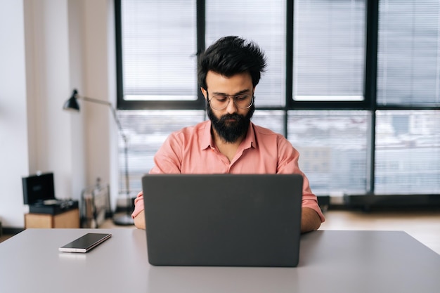 Retrato do homem de negócios indiano em óculos trabalhando no laptop sentado na mesa na sala de escritório de coworking leve pela janela