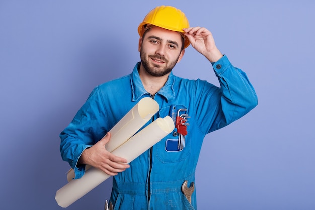 Retrato do jovem arquiteto barbudo atraente, construtor ou engenheiro, usando capacete amarelo e uniforme azul, capataz de pé com desenhos na mão e tocando seu capacete, olha para a câmera.