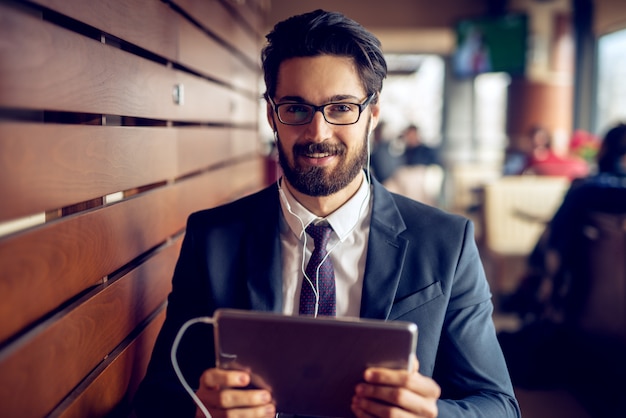 Retrato do jovem empresário barbudo bonito alegre em ouvir música de terno de um tablet em um café moderno e olhando para a câmera.