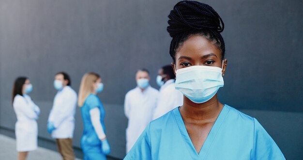 Retrato do jovem médico afro-americano de mulher bonita na máscara médica, olhando para a câmera. Perto da médica em proteção respiratória. Colegas de médicos de raças mistas no fundo.