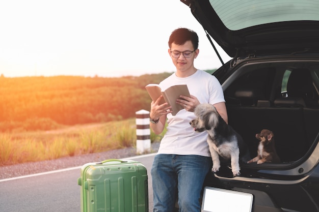 Foto retrato do livro de leitura asiático novo do homem ao sentar-se no tronco aberto do carro com seus cães.