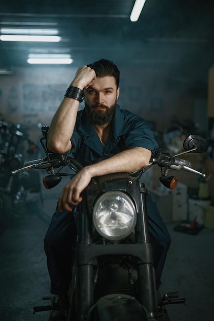 Retrato do mecânico de barba brutal homem sentado na motocicleta reparada sobre o fundo da oficina