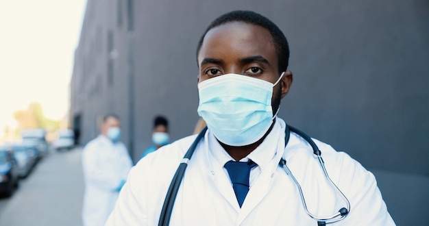 Retrato do médico afro-americano de homem bonito na máscara médica e com estetoscópio, olhando para a câmera. Feche de médico masculino na proteção respiratória. Médicos de raças mistas no fundo.