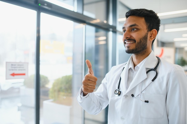 Retrato do médico indiano masculino vestindo jaleco branco com porta aberta no corredor da clínica como pano de fundo