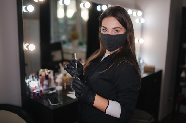 Foto retrato do mestre de sobrancelhas feminino profissional com manto preto com luvas pretas e máscara protetora preta usando pincel e henna para as sobrancelhas