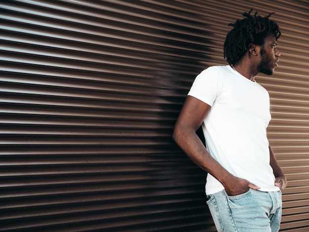 Retrato do modelo de hipster bonito sorridente Homem africano não barbeado vestido com camiseta branca de verão e jeans Moda masculina com penteado dreadlocks posando no fundo da rua