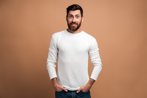 Retrato do modelo de jovem barbudo bonito brutal na camisa branca em pé e olhando para a câmera com rosto orgulhoso de prazer. estúdio interno tiro isolado em fundo bege