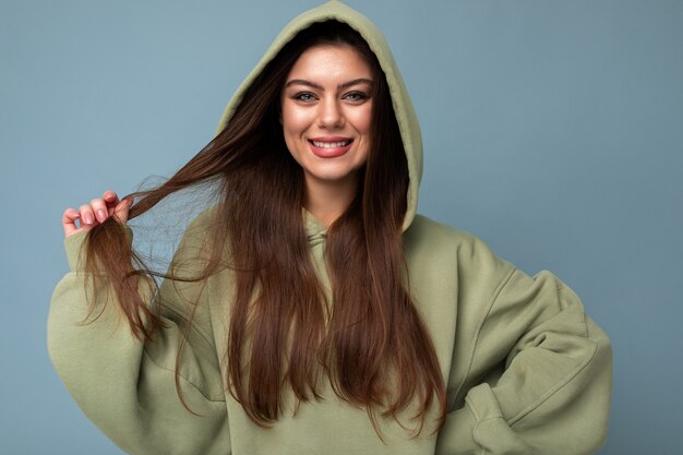 Retrato do modelo de mulher jovem e atraente caucasiano sorridente morena com capuz caqui moderno e capuz isolado sobre um fundo azul com espaço de cópia. Conceito positivo