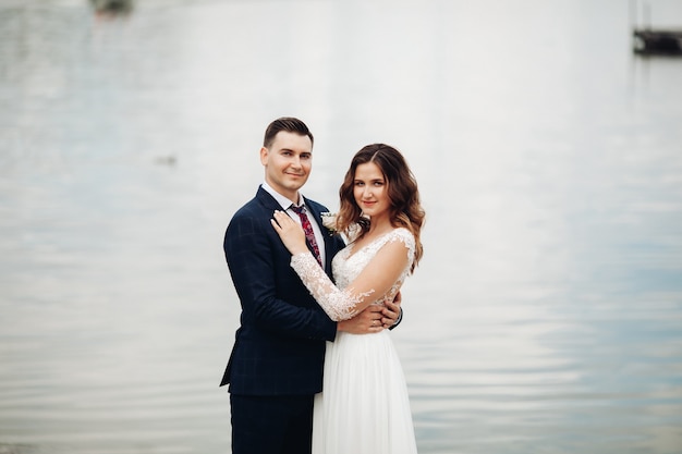 Retrato do noivo em um terno preto com uma linda noiva em um vestido branco no parque fica perto do lago