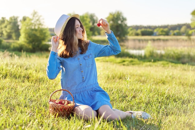 Retrato do país da bela adolescente com chapéu vestido jeans com cesta de morangos frescos, comer morango maduro. fundo de paisagem natural, prado verde, céu azul