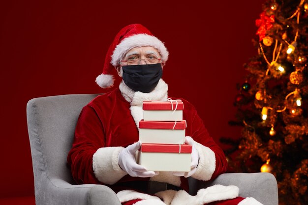 Retrato do papai noel em máscara protetora sentado na poltrona com caixas de presente ele preparando presentes para as crianças no natal