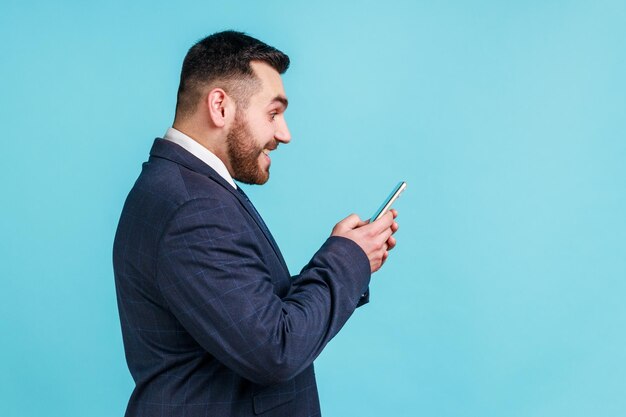 Foto retrato do perfil do empresário feliz vestindo terno estilo oficial em pé usando smartphone e mensagem de texto com sorriso verificando e-mail tiro de estúdio interno isolado em fundo azul