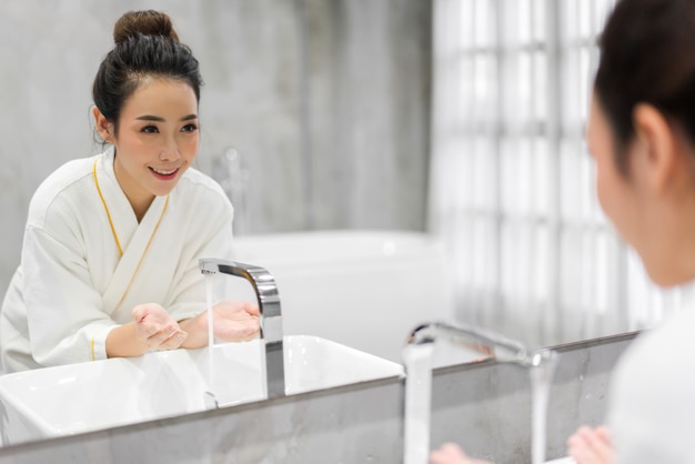 Retrato do sorriso da mulher asiática bonita alegre bonita pele branca saudável fresca limpa, lavar o rosto limpo com água na frente do espelho no banheiro. Beleza e spa. Pele fresca perfeita