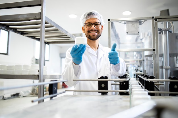 Retrato do tecnólogo em pé pela linha de montagem da fábrica de cosméticos e segurando a embalagem do creme