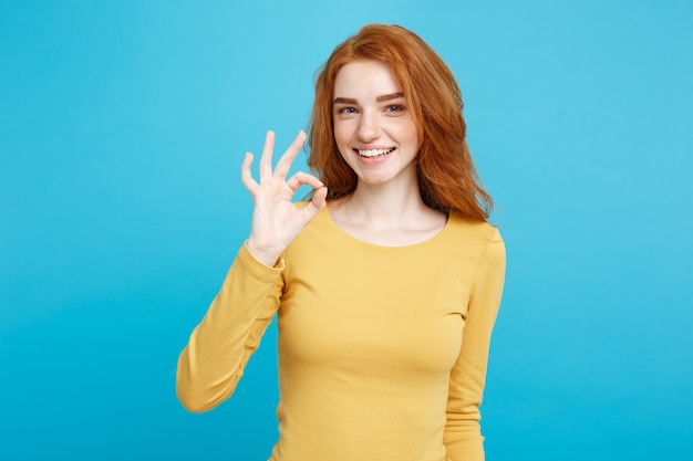 Retrato do tiro na cabeça do conceito de diversão e pessoas de uma encantadora garota ruiva ruiva com sardas sorrindo e fazendo sinal de ok com o dedo espaço na parede azul pastel