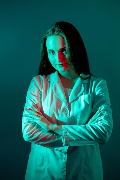 Retrato del doctor Profesional de la salud Carrera médica Confiada doctora en bata blanca de pie con los brazos cruzados en luz de neón roja aislada en azul verde azulado oscuro