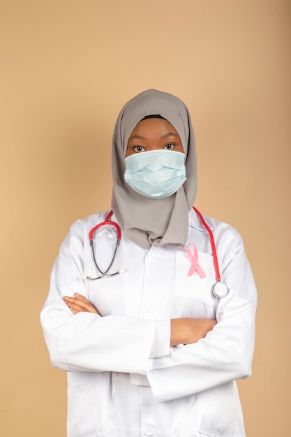 Retrato de una doctora afroamericana con una máscara quirúrgica, bufanda y bata blanca posando con los brazos cruzados.