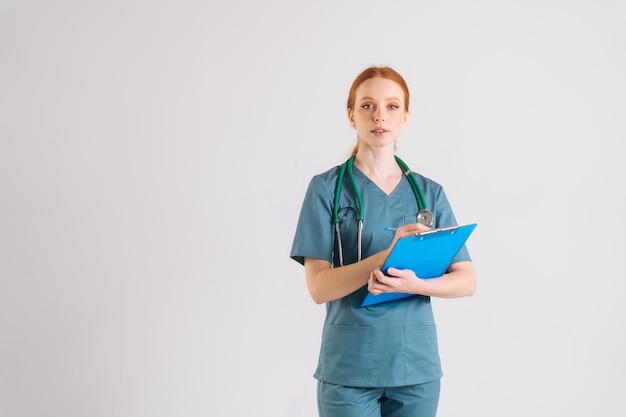 Retrato de una doctora atractiva con uniforme médico verde que escribe recetas en el portapapeles, completa el historial médico, mira la cámara, se para en un fondo blanco aislado en el estudio.