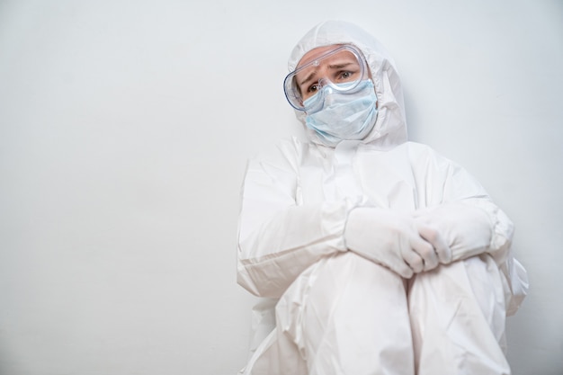 Retrato de una doctora cansada en un traje de protección, gafas, máscara y guante contra la pared blanca.