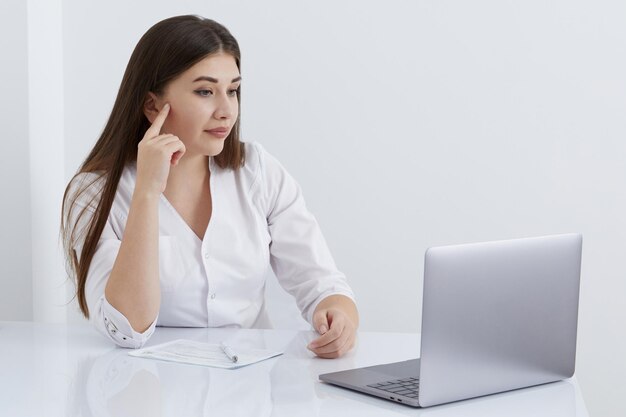 Retrato de una doctora hablando con una paciente en línea en la pantalla de una laptop sentada en el escritorio de la oficina de la clínica