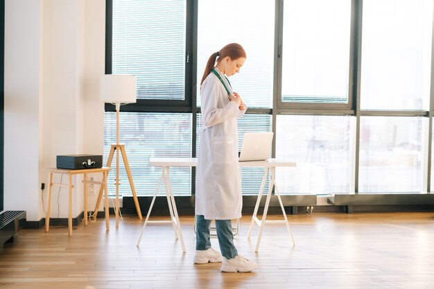 Retrato de una doctora pensativa en bata blanca de pie sobre el fondo de la ventana en un día soleado en la oficina de la clínica médica luz.