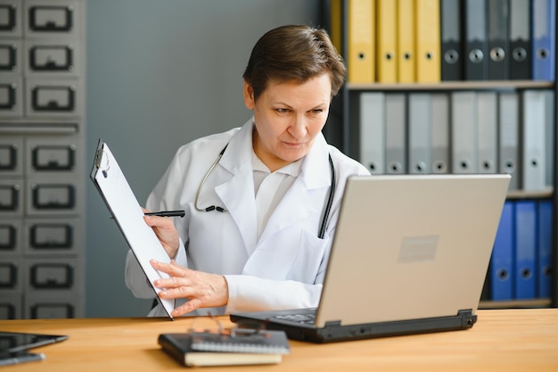 Retrato de una doctora que asesora a una paciente a través de una videollamada Médica profesional con bata blanca de laboratorio gesticulando y explicando el curso del tratamiento sentada en el escritorio de la oficina durante la consulta en línea