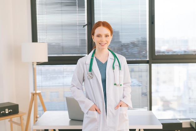 Retrato de una doctora sonriente con bata blanca parada en el fondo de la ventana en un día soleado en la oficina de una clínica médica ligera.